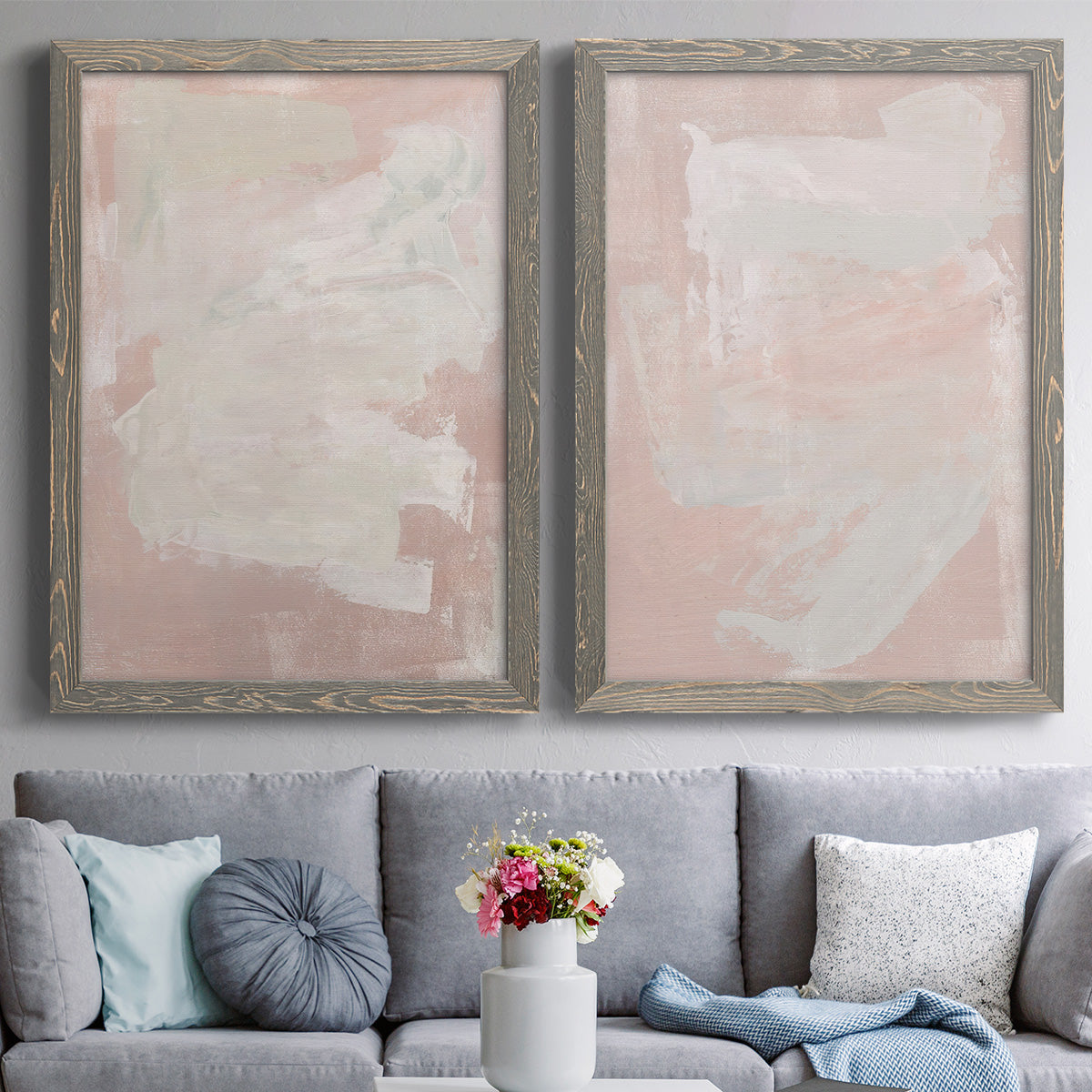 Set of two gray barnwood framed canvas prints depicting paired minimalist color-field abstracts with cloudy rose washes, broad pale strokes, and subtle texture above a sofa in a contemporary living room.