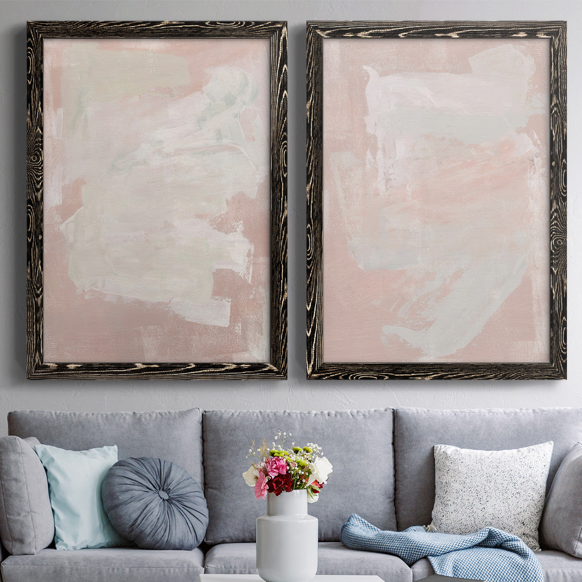 Set of two black barnwood framed canvas prints depicting paired minimalist color-field abstracts with cloudy rose washes, broad pale strokes, and subtle texture above a sofa in a contemporary living room.
