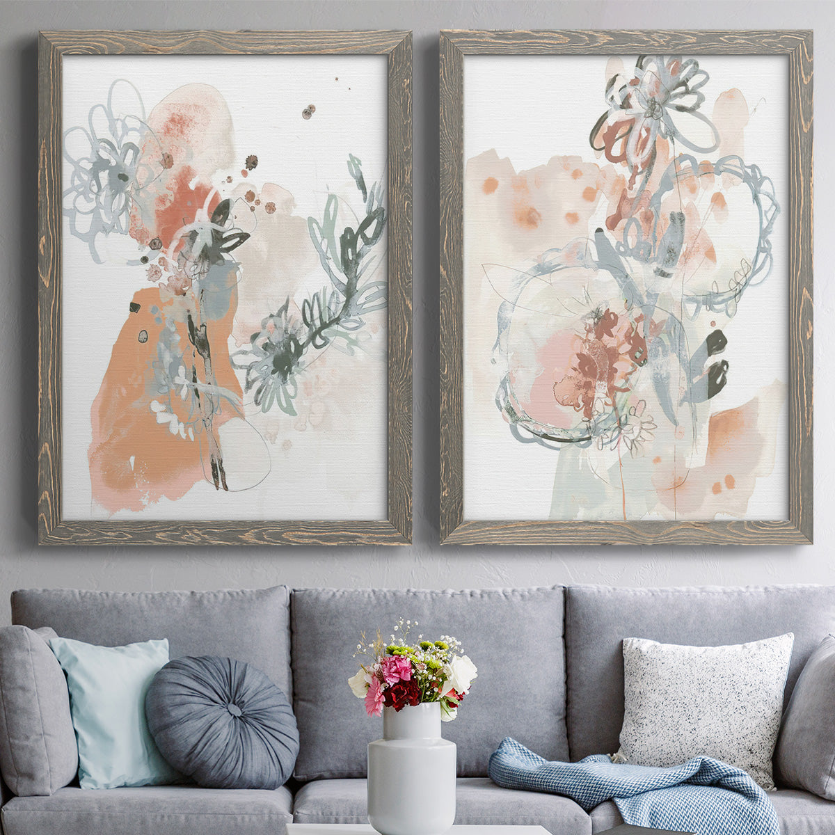 Set of two gray barnwood framed canvas prints depicting paired abstract watercolor botanicals with scribbled petals, translucent washes, and drifting stems above a sofa in a contemporary living room.