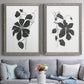 Set of two gray barnwood framed canvas prints depicting paired minimalist botanical prints with bold dark leaves, curving stems, and clean white grounds above a sofa in a contemporary living room.