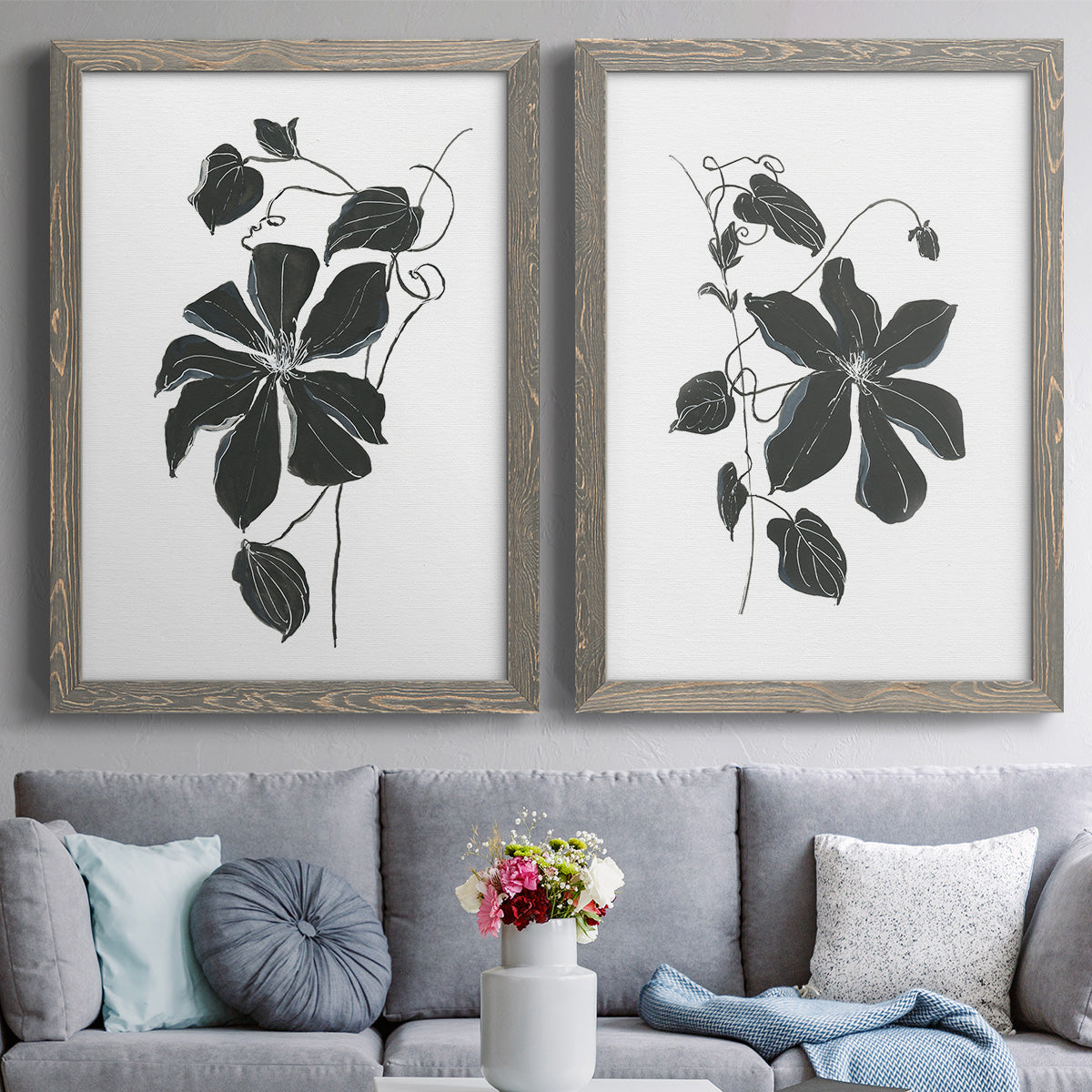 Set of two gray barnwood framed canvas prints depicting paired minimalist botanical prints with bold dark leaves, curving stems, and clean white grounds above a sofa in a contemporary living room.