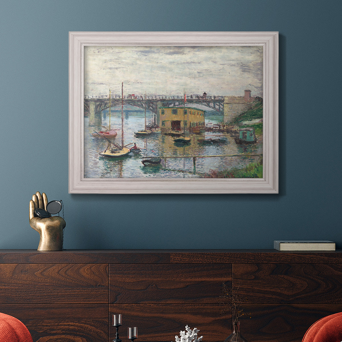 Impressionist river bridge scene on an overcast day by Claude Monet in an ornate weathered white frame displayed in a dark-toned interior setting.