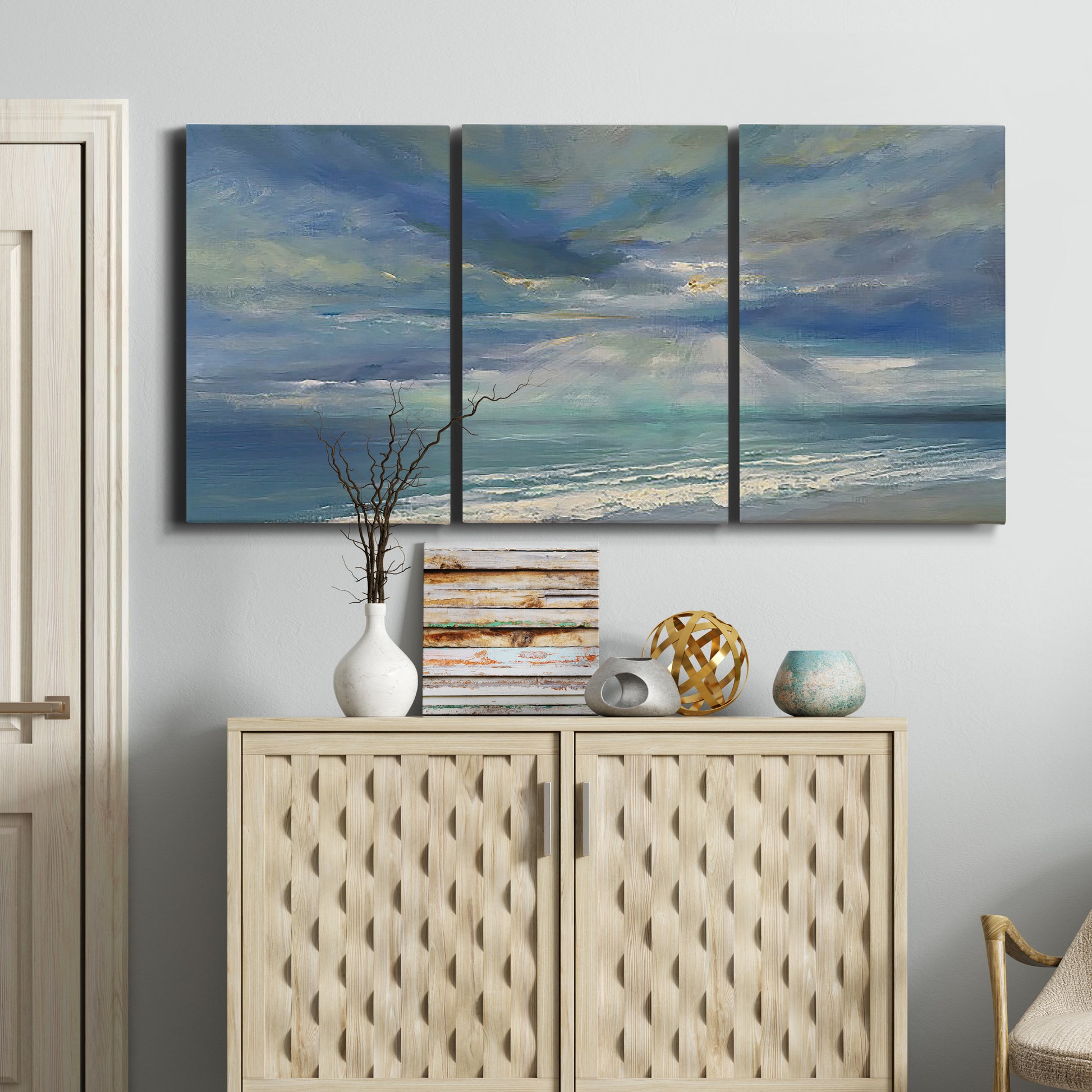 Three-panel canvas wall art featuring sunbeams breaking through coastal clouds, displayed above a cabinet in an entryway.