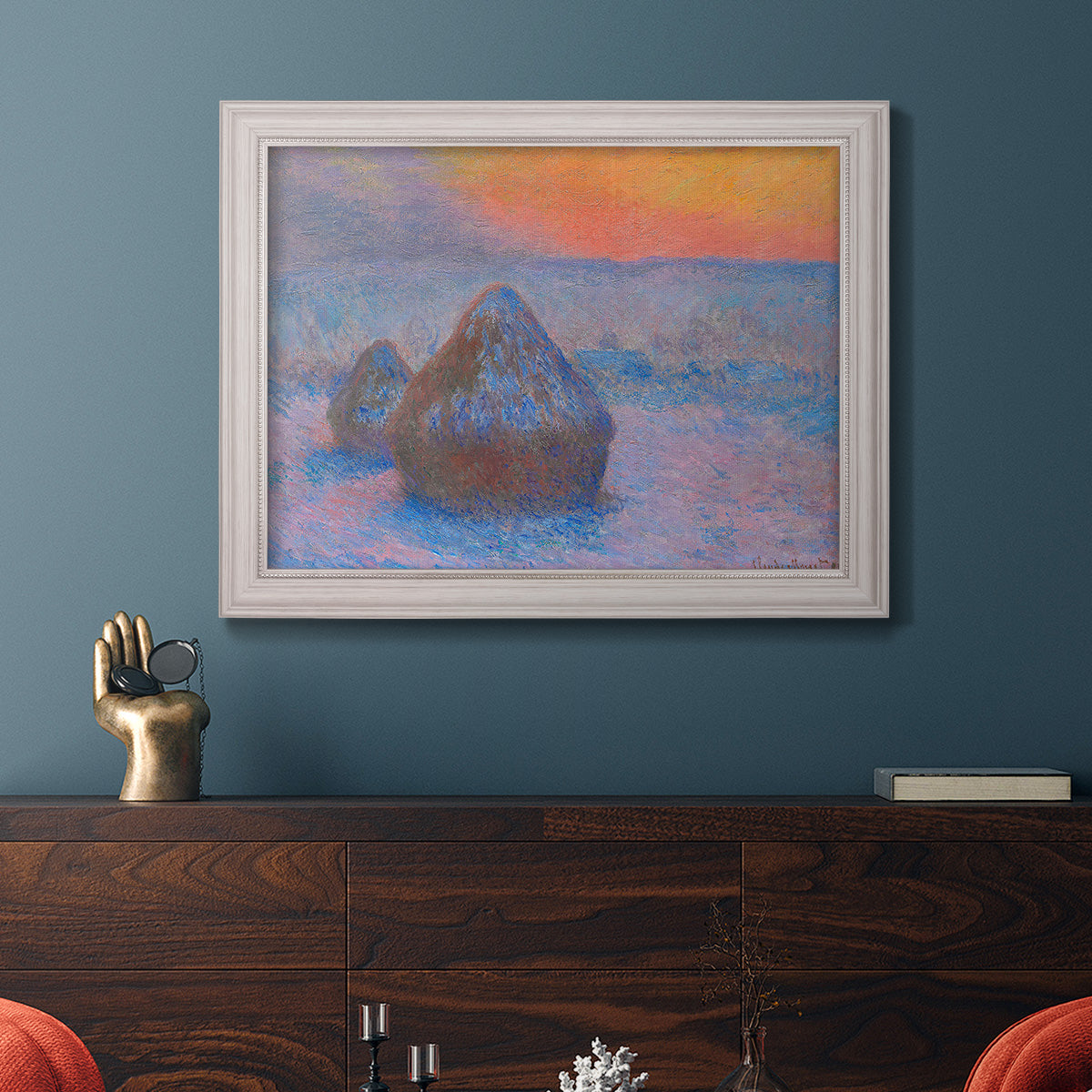 Impressionist haystack scene at sunset by Claude Monet in an ornate weathered white frame displayed in a dark-toned interior setting.