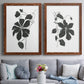 Set of two maple barnwood framed canvas prints depicting paired minimalist botanical prints with bold dark leaves, curving stems, and clean white grounds above a sofa in a contemporary living room.
