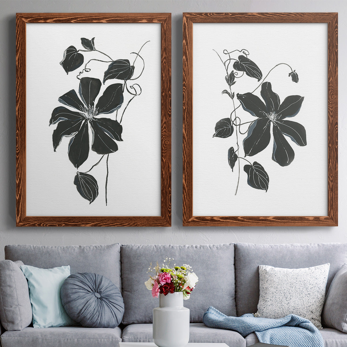 Set of two maple barnwood framed canvas prints depicting paired minimalist botanical prints with bold dark leaves, curving stems, and clean white grounds above a sofa in a contemporary living room.