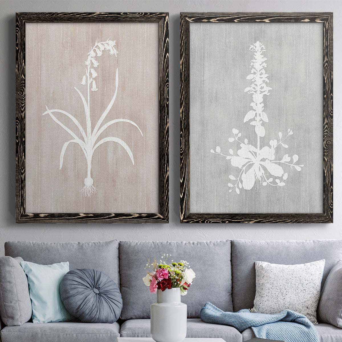 Set of two black barnwood framed canvas prints depicting paired rustic minimalist botanical prints with delicate stems, leaves, and blossoms against gray taupe and pale blue panels above a sofa in a contemporary living room.