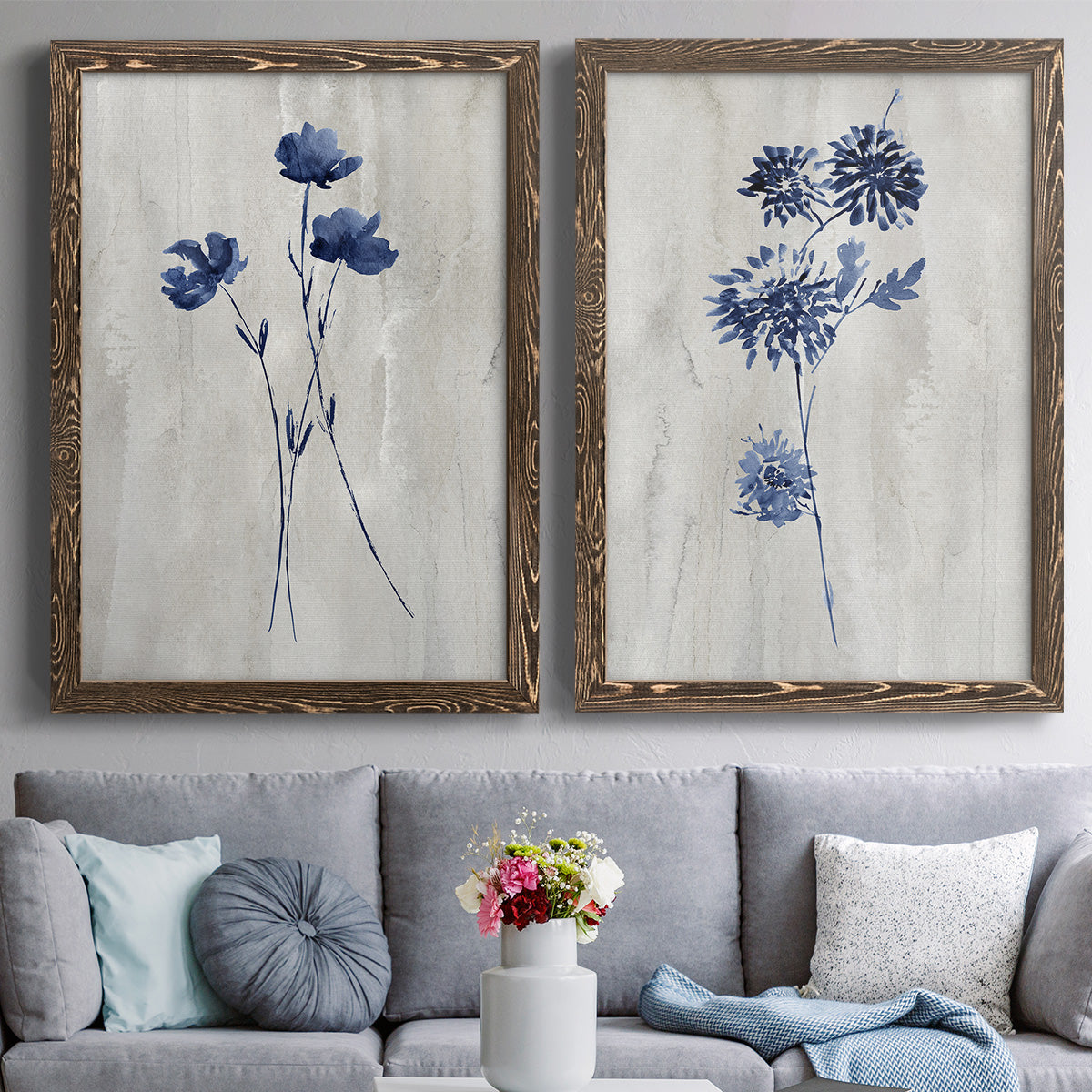 Set of two walnut barnwood framed canvas prints depicting paired rustic monochrome botanical paintings with indigo blossoms, thin stems, and softly distressed gray backgrounds above a sofa in a contemporary living room.