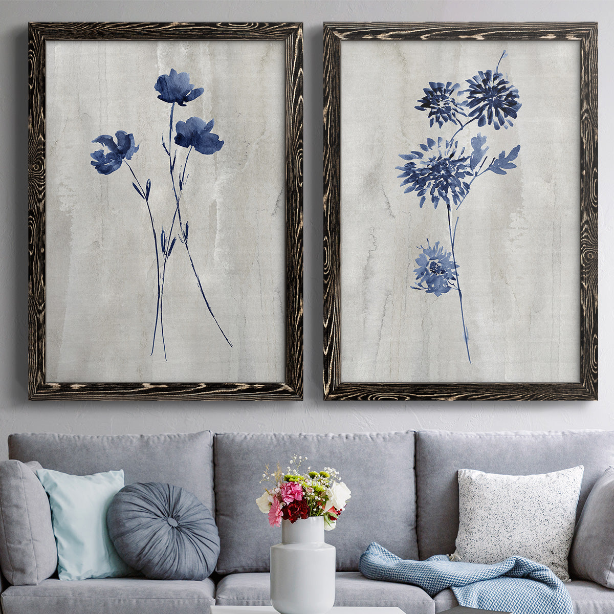 Set of two black barnwood framed canvas prints depicting paired rustic monochrome botanical paintings with indigo blossoms, thin stems, and softly distressed gray backgrounds above a sofa in a contemporary living room.