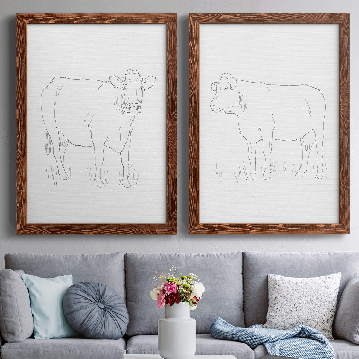 Set of two maple barnwood framed canvas prints depicting paired minimalist livestock sketches with fine gray lines, white backgrounds, and quiet farm-animal profiles above a sofa in a contemporary living room.