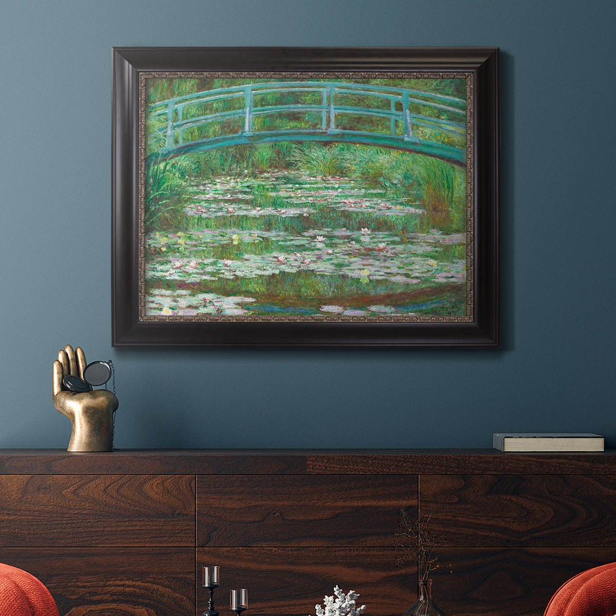 Impressionist garden scene with Japanese bridge and water lilies by Claude Monet in an ornate black frame with gilded inner detail displayed in a dark-toned interior setting.