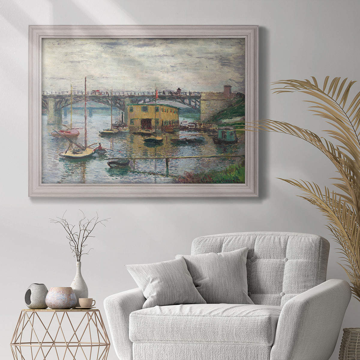 Impressionist river bridge scene on an overcast day by Claude Monet in an ornate weathered white frame displayed in a bright interior setting.