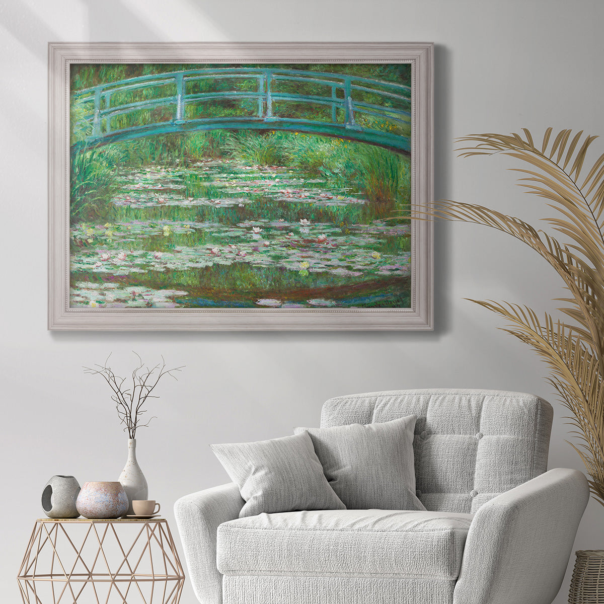 Impressionist garden scene with Japanese bridge and water lilies by Claude Monet in an ornate weathered white frame displayed in a bright interior setting.