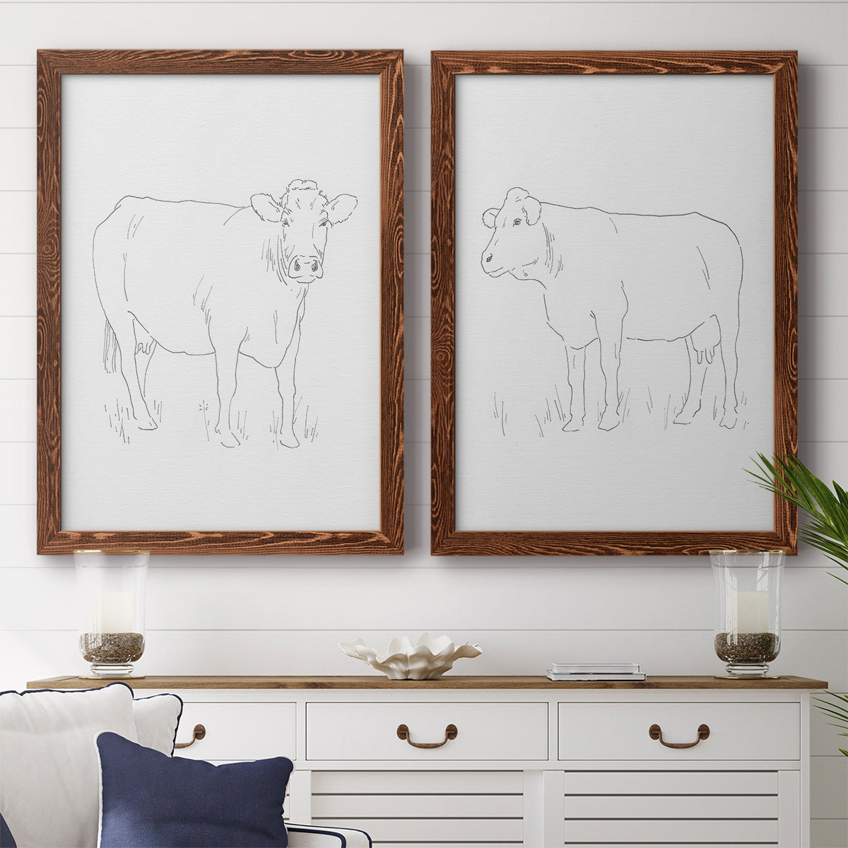 Set of two maple barnwood framed canvas prints depicting a coordinated art pair of simple contour drawings of standing cows rendered as minimalist livestock sketches above a dining room sideboard.
