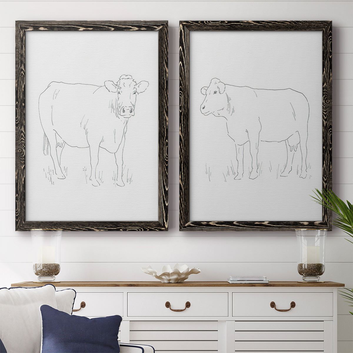 Set of two black barnwood framed canvas prints depicting a coordinated art pair of simple contour drawings of standing cows rendered as minimalist livestock sketches above a dining room sideboard.