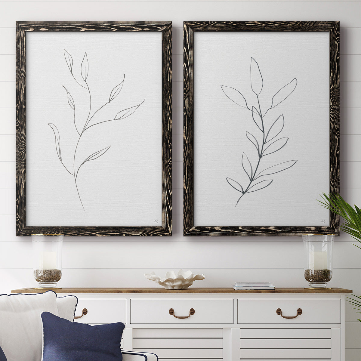 Set of two black barnwood framed canvas prints depicting a coordinated art pair of single continuous-line leafy branches rendered as minimalist botanical line drawings above a dining room sideboard.