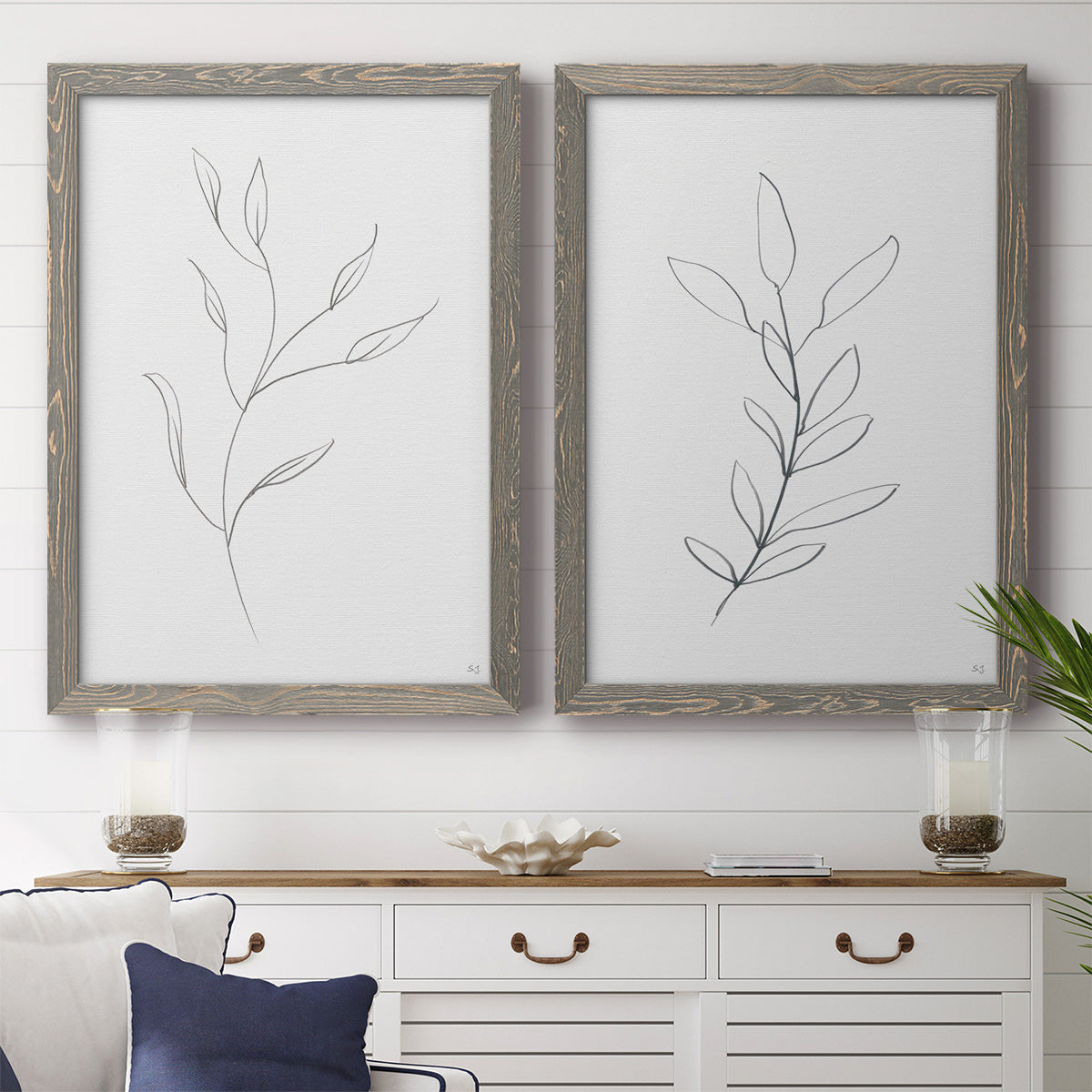 Set of two gray barnwood framed canvas prints depicting a coordinated art pair of single continuous-line leafy branches rendered as minimalist botanical line drawings above a dining room sideboard.