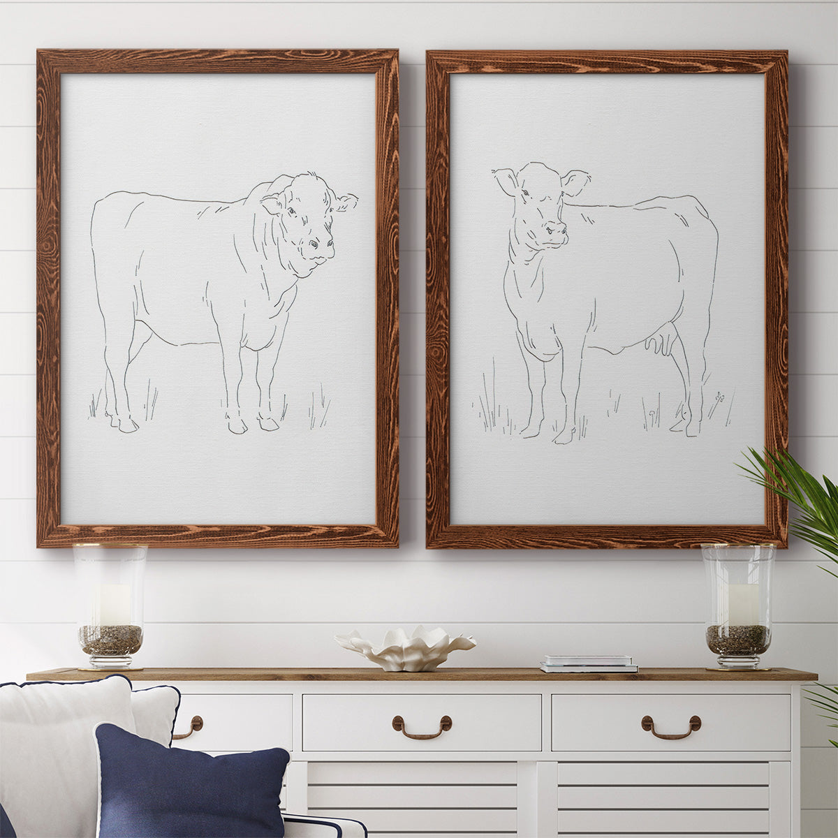 Set of two maple barnwood framed canvas prints depicting a coordinated art pair of simple contour drawings of standing cows rendered as minimalist livestock sketches above a dining room sideboard.