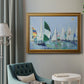 Coastal harbor watercolor painting in an ornate traditional gold frame displayed in a neutral interior setting.