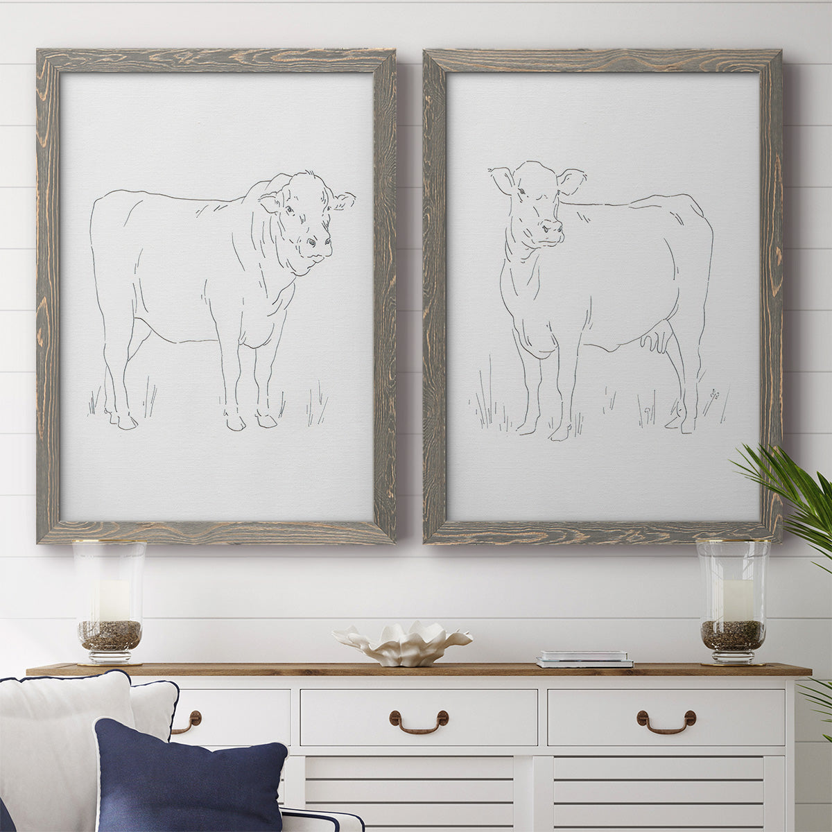 Set of two gray barnwood framed canvas prints depicting a coordinated art pair of simple contour drawings of standing cows rendered as minimalist livestock sketches above a dining room sideboard.