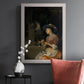 Classical interior scene of a woman weaving flowers by Godfried Schalcken in an ornate weathered white frame displayed in a dark-toned interior setting.