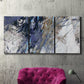 Three-panel canvas wall art featuring textured blue abstract composition, displayed in a living room above a suede accent chair.