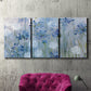 Three-panel canvas wall art featuring delicate watercolor floral blooms, displayed in a living room above a suede accent chair.