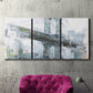 Three-panel canvas wall art featuring urban cityscape with architectural forms, displayed in a living room above a suede accent chair.