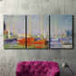 Three-panel canvas wall art featuring red boats along riverbank, displayed in a living room above a suede accent chair.