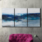 Three-panel canvas wall art featuring moody abstract sea with depth, displayed in a living room above a suede accent chair.