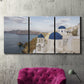 Three-panel canvas wall art featuring white buildings overlooking sea cliffs, displayed in a living room above a suede accent chair.