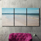 Three-panel canvas wall art featuring white sand beach with gentle waves, displayed in a living room above a suede accent chair.