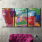 Three-panel canvas wall art featuring vibrant abstract color field panels, displayed in a living room above a suede accent chair.