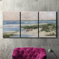Three-panel canvas wall art featuring soft morning light over calm sea, displayed in a living room above a suede accent chair.