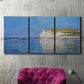 Three-panel canvas wall art featuring white cliffs rising above sea, displayed in a living room above a suede accent chair.