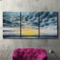 Three-panel canvas wall art featuring tropical coastal sunset over water, displayed in a living room above a suede accent chair.