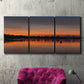 Three-panel canvas wall art featuring glowing sunset over water, displayed in a living room above a suede accent chair.