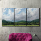 Three-panel canvas wall art featuring pastoral mountain landscape in blue, displayed in a living room above a suede accent chair.