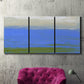 Three-panel canvas wall art featuring abstract coastal inlet landscape, displayed in a living room above a suede accent chair.