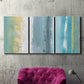 Three-panel canvas wall art featuring abstract coastal beach textures, displayed in a living room above a suede accent chair.