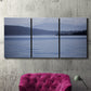 Three-panel canvas wall art featuring calm blue water landscape, displayed in a living room above a suede accent chair.