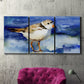 Three-panel canvas wall art featuring shorebirds standing in shallow water, displayed in a living room above a suede accent chair.