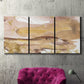 Three-panel canvas wall art featuring layered abstract dawn tones, displayed in a living room above a suede accent chair.