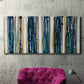 Three-panel canvas wall art featuring vertical blue abstract streaks, displayed in a living room above a suede accent chair.