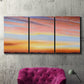 Three-panel canvas wall art featuring glowing sunset sky over landscape, displayed in a living room above a suede accent chair.