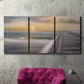 Three-panel canvas wall art featuring tropical beach at sunset photograph, displayed in a living room above a suede accent chair.