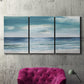 Three-panel canvas wall art featuring calm coastal shoreline, displayed in a living room above a suede accent chair.