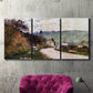 Three-panel canvas wall art featuring country road through village, displayed in a living room above a suede accent chair.