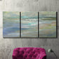 Three-panel canvas wall art featuring soft coastal beach light landscape, displayed in a living room above a suede accent chair.
