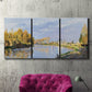 Three-panel canvas wall art featuring river flowing past village, displayed in a living room above a suede accent chair.
